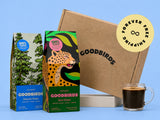 Two GoodBirds boxes, one blue with trees illustration and one green with a leopard illustration, next to a large brown cardboard shipping box and a glass of black coffee. A yellow circular badge reads 'Forever Free Shipping'.