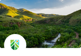 A scenic landscape photo of rolling green hills, a winding river, and a blue sky, with a white circular overlay in the lower left containing a green and blue shield-like logo.